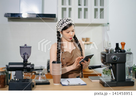 Focused Asian female barista taking stock and managing inventory with a clipboard at a modern cafe counter. Focused Asian female barista taking stock and managing inventory with a clipboard at a modern cafe counter. 141458951