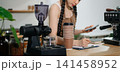 Focused Asian female barista taking stock and managing inventory with a clipboard at a modern cafe counter. 141458952