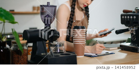 Focused Asian female barista taking stock and managing inventory with a clipboard at a modern cafe counter. Focused Asian female barista taking stock and managing inventory with a clipboard at a modern cafe counter. 141458952