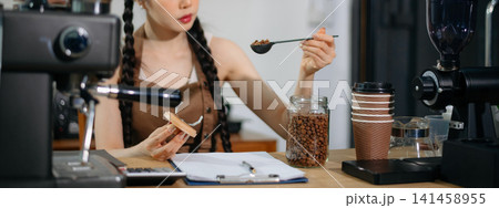 Young Asian female barista preparing fresh roasted coffee beans in a modern cafe kitchen with professional espresso equipment. Young Asian female barista preparing fresh roasted coffee beans in a modern cafe kitchen with professional espresso equipment. 141458955
