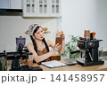 Young Asian female barista preparing fresh roasted coffee beans in a modern cafe kitchen with professional espresso equipment. 141458957