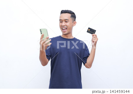 Man smiles broadly, holding phone and credit card. He appears excited, possibly about a purchase or payment. Simple background highlights his joyful expression. Image suits ecommerce Man smiles broadly, holding phone and credit card. He appears excited, possibly about a purchase or payment. Simple background highlights his joyful expression. Image suits ecommerce 141459144