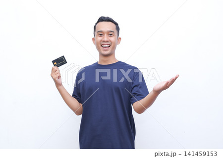 Young man smiles, holding a credit card. He wears a dark blue tshirt. His posture shows confidence and ease. The white background is clean and simple. Ideal for financial, shopping 141459153