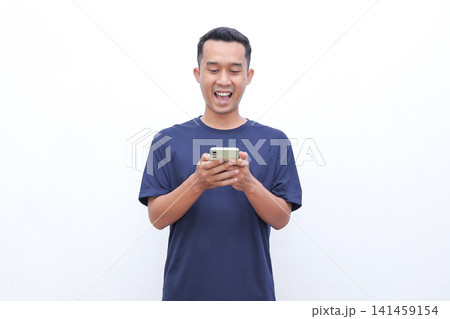 Man smiles, holding phone with excited expression. His eyes widen, mouth opens in joyful surprise. Fingers tap screen as he reacts to content. Ideal for online shopping, social media 141459154