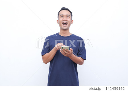 Young man holds phone with wide eyed surprise. He stares ahead, mouth open in astonishment. Simple white background highlights his reaction. Perfect for digital surprises, unexpected news 141459162