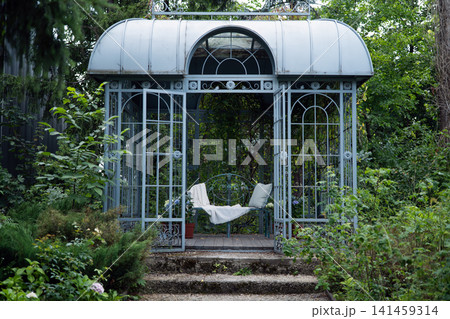 Vintage metal gazebo with open doors and bench in lush green garden, summer outdoor landscape. Vintage metal gazebo with open doors and bench in lush green garden, summer outdoor landscape. 141459314