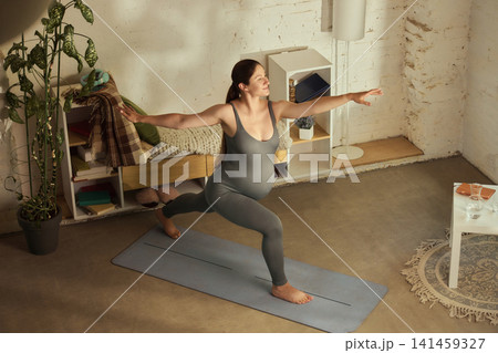 Focused athletic pregnant brunette practicing warrior yoga pose at home 141459327