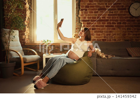 Pregnant woman relaxing on beanbag and using smartphone at home 141459342
