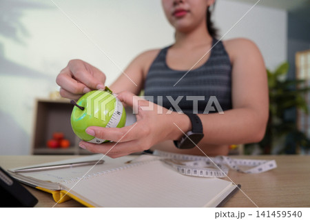 Woman wrapping measuring tape around green apple for weight loss and nutrition planning Woman wrapping measuring tape around green apple for weight loss and nutrition planning 141459540