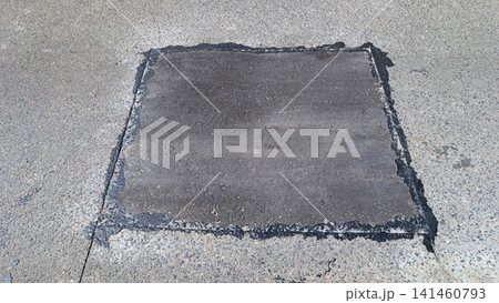 Rectangular fresh asphalt repair patch on light concrete background. Dark gray rough texture with wet sheen along edges. Natural daylight highlights contrast between old new pavement Rectangular fresh asphalt repair patch on light concrete background. Dark gray rough texture with wet sheen along edges. Natural daylight highlights contrast between old new pavement 141460793
