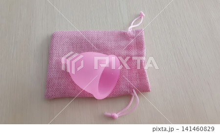Pink menstrual cup resting on a textured pink drawstring pouch on a light wooden surface, top down view. Pink menstrual cup resting on a textured pink drawstring pouch on a light wooden surface, top down view. 141460829