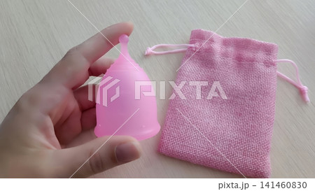 Hand holding a translucent pink menstrual cup beside a pink drawstring pouch on a light wooden surface. Hand holding a translucent pink menstrual cup beside a pink drawstring pouch on a light wooden surface. 141460830