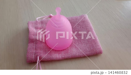 Translucent pink menstrual cup with ridged top and pull tab, resting on a pink drawstring fabric pouch on wood. Translucent pink menstrual cup with ridged top and pull tab, resting on a pink drawstring fabric pouch on wood. 141460832