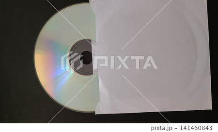 Close up of a CD with holographic sheen partially covered by a white paper sleeve with a semi circular cutout, on a dark background. Close up of a CD with holographic sheen partially covered by a white paper sleeve with a semi circular cutout, on a dark background. 141460843