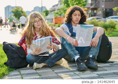 Two teenage students sitting outdoors reading workbook preparing for exams together 141462645