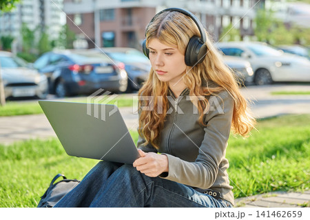 Young woman in headphones working on laptop outdoors sitting on green grass Young woman in headphones working on laptop outdoors sitting on green grass 141462659