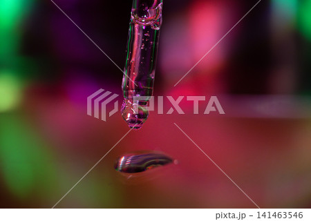 Close-up of a dropper releasing a drop of purple liquid onto a reflective surface, with colorful bokeh lights creating a vibrant background Close-up of a dropper releasing a drop of purple liquid onto a reflective surface, with colorful bokeh lights creating a vibrant background 141463546