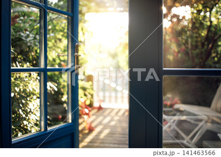 Blue-framed glass door open to a sunlit garden with wooden deck, greenery, and a lounge chair visible in the background creating a serene outdoor space 141463566