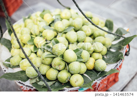 Fresh green citrus fruits arranged in a woven basket with leaves, suspended by ropes, placed on a textured surface in a market setting 141463574
