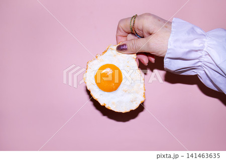 Hand holding a fried egg with a bright yellow yolk against a pastel pink background, showcasing a vibrant and playful food presentation Hand holding a fried egg with a bright yellow yolk against a pastel pink background, showcasing a vibrant and playful food presentation 141463635