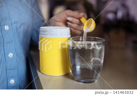 Person in blue shirt adds yellow powder supplement to clear glass of water on wooden table, container with yellow lid visible in the background Person in blue shirt adds yellow powder supplement to clear glass of water on wooden table, container with yellow lid visible in the background 141463832