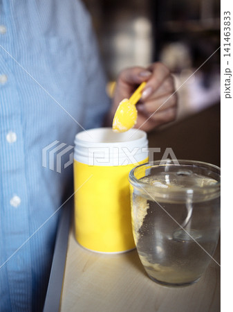 Person in blue shirt using yellow spoon to scoop powder from yellow container into clear glass of water on wooden surface in a kitchen setting Person in blue shirt using yellow spoon to scoop powder from yellow container into clear glass of water on wooden surface in a kitchen setting 141463833