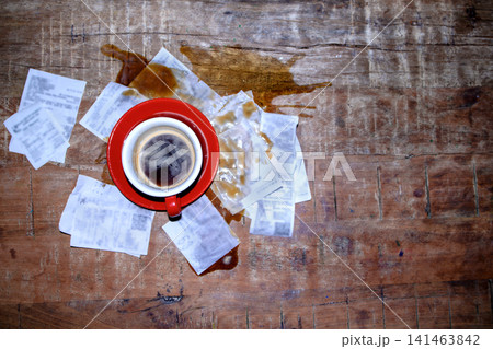 Red coffee cup filled with black coffee placed on a wooden table surrounded by spilled coffee and crumpled receipts scattered around Red coffee cup filled with black coffee placed on a wooden table surrounded by spilled coffee and crumpled receipts scattered around 141463842