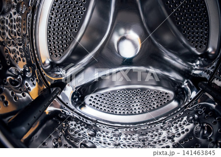 Close-up view of the interior of a stainless steel washing machine drum showcasing perforated holes and water droplets on the surface Close-up view of the interior of a stainless steel washing machine drum showcasing perforated holes and water droplets on the surface 141463845
