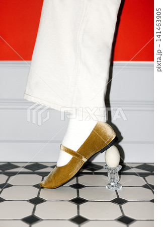 Female foot in yellow velvet shoe resting on a glass egg holder with an egg, standing on a black and white tiled floor against a red wall Female foot in yellow velvet shoe resting on a glass egg holder with an egg, standing on a black and white tiled floor against a red wall 141463905