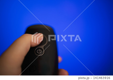 Hand holding a black remote control with a power button against a bright blue background, emphasizing the device's design and functionality in a modern setting Hand holding a black remote control with a power button against a bright blue background, emphasizing the device's design and functionality in a modern setting 141463916