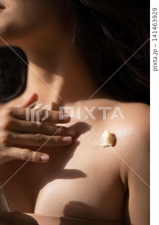 Close-up of a woman applying body cream on her shoulder, with soft lighting highlighting the skin texture and a small amount of cream visible on the skin Close-up of a woman applying body cream on her shoulder, with soft lighting highlighting the skin texture and a small amount of cream visible on the skin 141463929