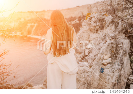 Woman cliff sunset standing on a rocky coastal ledge looking at the ocean with long flowing hair during golden hour Woman cliff sunset standing on a rocky coastal ledge looking at the ocean with long flowing hair during golden hour 141464246