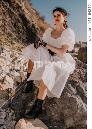 Woman white dress coast sitting elegantly on rugged seaside stones during golden hour for a lifestyle fashion portrait Woman white dress coast sitting elegantly on rugged seaside stones during golden hour for a lifestyle fashion portrait 141464250