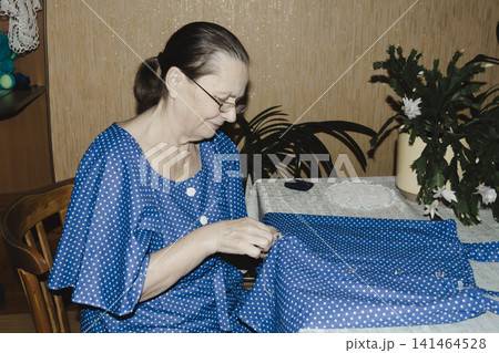 An elderly woman in blue dress focuses intently sewing garment table quiet domestic scene highlighting craftsmanship, patience, and the timeless joy creating clothing hand cozy familiar home setting 141464528