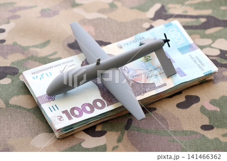 Unnamed grey toy drone and ukrainian money bills on camouflage fabric close up 141466362