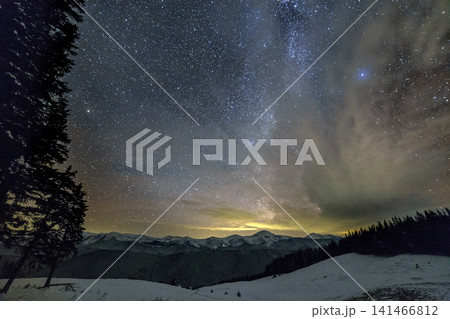 Mountain winter night panorama after sunset. Steep long ridge mountain peaks, deep snow in valley, hills covered with dense spruce forest, dramatic snowy stormy cloud on dark blue starry sky. 141466812