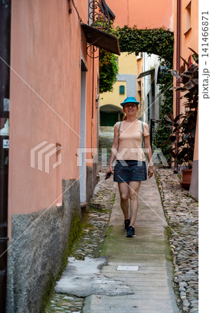 Cervo, liguria, italy. June 22, 2025. Woman in a blue hat walking through a picturesque cobblestone alley in the historic medieval village of cervo, liguria, italy Cervo, liguria, italy. June 22, 2025. Woman in a blue hat walking through a picturesque cobblestone alley in the historic medieval village of cervo, liguria, italy 141466871