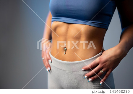Fit woman showing defined abs in blue sports top and gray leggings with belly button piercing 141468259