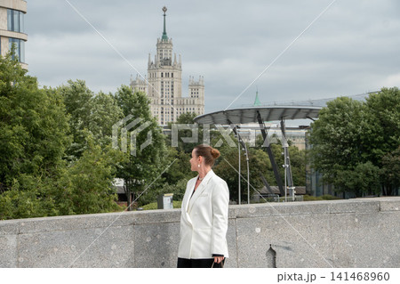 Woman stands in Zaryadye Park with Kotelnicheskaya Embankment Building and modern park architecture in background on cloudy day. Urban lifestyle, travel, sightseeing, city landmark, outdoor recreation 141468960