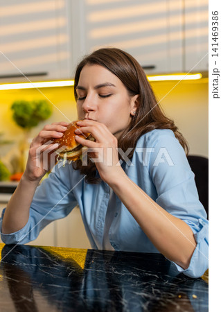 Young woman enjoying tasty burger in home kitchen indoors satisfied with quick fast food cheat lunch Young woman enjoying tasty burger in home kitchen indoors satisfied with quick fast food cheat lunch 141469386