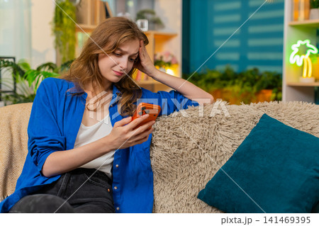 Young woman on home sofa feeling upset after bad news on smartphone while taking heavy sigh indoors 141469395