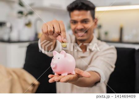 Indian man drops coins into piggy bank listens to ring sound smiles plans savings invest for future Indian man drops coins into piggy bank listens to ring sound smiles plans savings invest for future 141469409