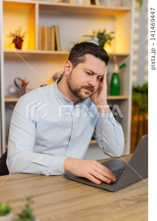Young man on home table with headache rubbing temples while working on laptop after burnout indoors Young man on home table with headache rubbing temples while working on laptop after burnout indoors 141469447