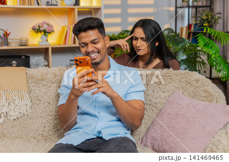Indian couple man and woman sitting on home sofa checking smartphone messages with surprise reaction 141469465