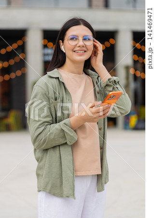 Young woman studying with audio lesson walking in city street with earbuds and glasses like student Young woman studying with audio lesson walking in city street with earbuds and glasses like student 141469471
