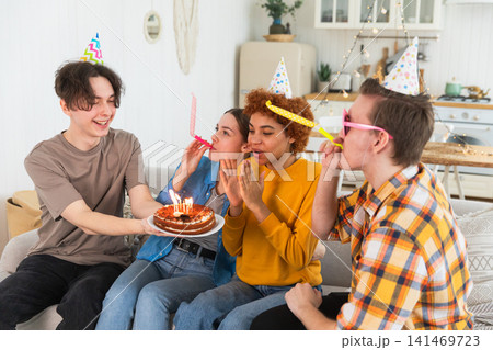 Make a wish. Woman wearing party cap blowing out burning candles on birthday cake. Happy Birthday party. Group of friends wishes girl happy birthday. People celebrating birthday with party at home Make a wish. Woman wearing party cap blowing out burning candles on birthday cake. Happy Birthday party. Group of friends wishes girl happy birthday. People celebrating birthday with party at home 141469723