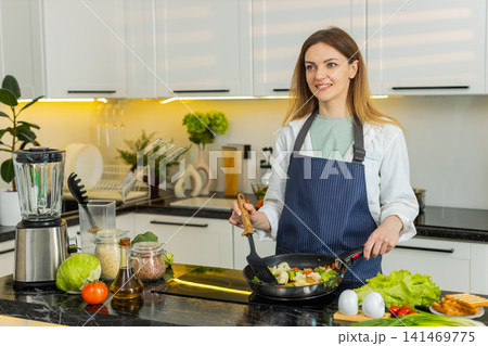 Middle-aged woman cooking fresh vegetables in frying pan in home kitchen for healthy diet meal plan 141469775