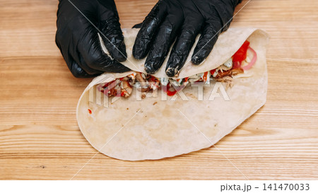 Pita sandwich. Preparing burrito. Chef cooking shawarma on kitchen table. Gloved hands wrapping flatbread with chicken vegetables sauce. Pita sandwich. Preparing burrito. Chef cooking shawarma on kitchen table. Gloved hands wrapping flatbread with chicken vegetables sauce. 141470033