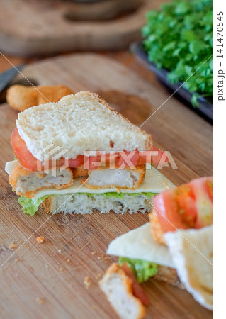 Homemade crispy chicken sandwich layered with fresh tomato, cheese, and lettuce on artisan bread. Rustic lunch, meal preparation, cafe food, and healthy homemade cooking concept. 141470545