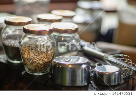 Glass jars filled with oats and baking ingredients alongside measuring cups and a whisk on a rustic wooden surface. Home baking, recipe preparation, artisan kitchen, and healthy cooking concept. Glass jars filled with oats and baking ingredients alongside measuring cups and a whisk on a rustic wooden surface. Home baking, recipe preparation, artisan kitchen, and healthy cooking concept. 141470551
